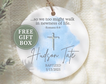 May include: A white ceramic ornament with a gold string for hanging. The ornament features the text "...so we too might walk in newness of life." and "Hudson Tate BAPTIZED 5/13/2025". A cross design is centered above the name.