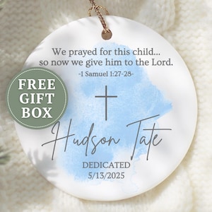 May include: A white ceramic ornament with a gold string for hanging. The ornament features the text "We prayed for this child..." and "Hudson Tate" with a cross and a blue watercolor design. Includes the text "Dedicated 5/13/2025" and a "FREE GIFT BOX" sticker.