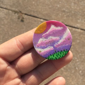 May include: A round, painted pin with a purple, pink, and yellow sunset scene with green trees at the bottom. The pin has a glittery texture.