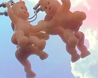 Op de afbeelding: Een paar zilveren oorbellen met kleine, gedetailleerde plastic babyfiguurtjes. De baby's staan in een speelse pose, waarbij de een de hand van de ander vasthoudt. De oorbellen zijn een grillige en unieke accessoire.