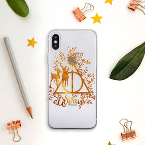 Puede incluir: Una funda transparente para iPhone con un diseño dorado que presenta el símbolo de las Reliquias de la Muerte, dos ciervos dorados y la palabra "Always" con la palabra "iPhone" debajo.