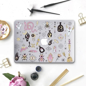 May include: A gray laptop with a decorative design featuring mystical symbols like a snake, moon, stars, and insects. The design includes the word "MAGIC" on a banner, along with other esoteric illustrations. The laptop is surrounded by stationery and decorative items.