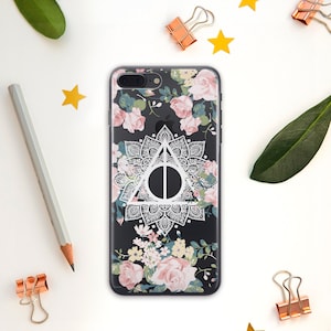 May include: A clear phone case with a black background and a white floral pattern. The case features a white mandala design with a white triangle and circle in the center. The triangle and circle are the Deathly Hallows symbol from the Harry Potter series.