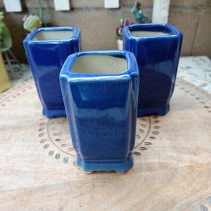 May include: Three square-shaped ceramic vessels with a glossy, deep blue crackle glaze finish. Each container features a slightly flared rim, a smooth interior, and a small, raised base. The vessels are arranged in a triangular formation.