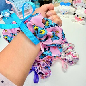 May include: A collection of colorful scrunchies. One pink scrunchie features a blue cartoon character print and a blue ribbon. Other scrunchies have floral and character prints. The scrunchies are made of a soft, silky material.
