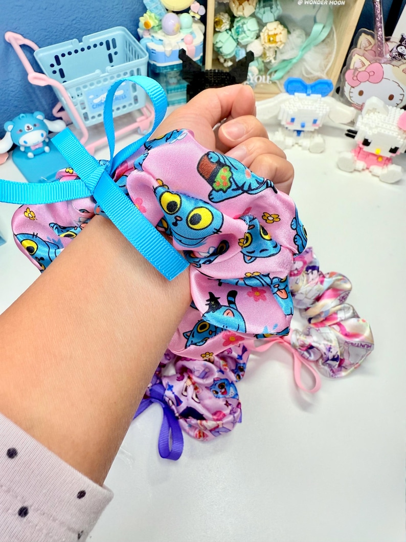 May include: A collection of colorful scrunchies. One pink scrunchie features a blue cartoon character print and a blue ribbon. Other scrunchies have floral and character prints. The scrunchies are made of a soft, silky material.