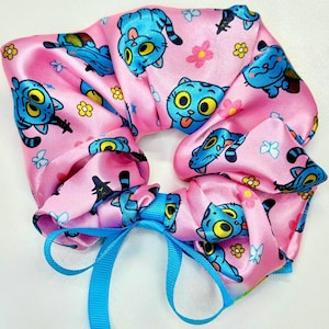 May include: Pink satin scrunchie with a repeating pattern of blue cartoon cats, yellow flowers, and butterflies. A blue ribbon is tied in a bow at the base of the scrunchie. The scrunchie is designed for hair.