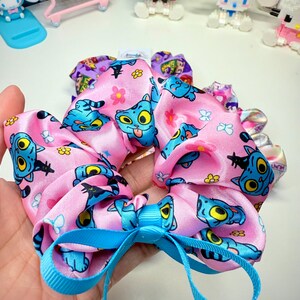 May include: Pink satin scrunchie with a repeating cartoon cat pattern in blue and yellow. A light blue ribbon is tied in a bow at the center. Other scrunchies with different patterns are visible in the background.