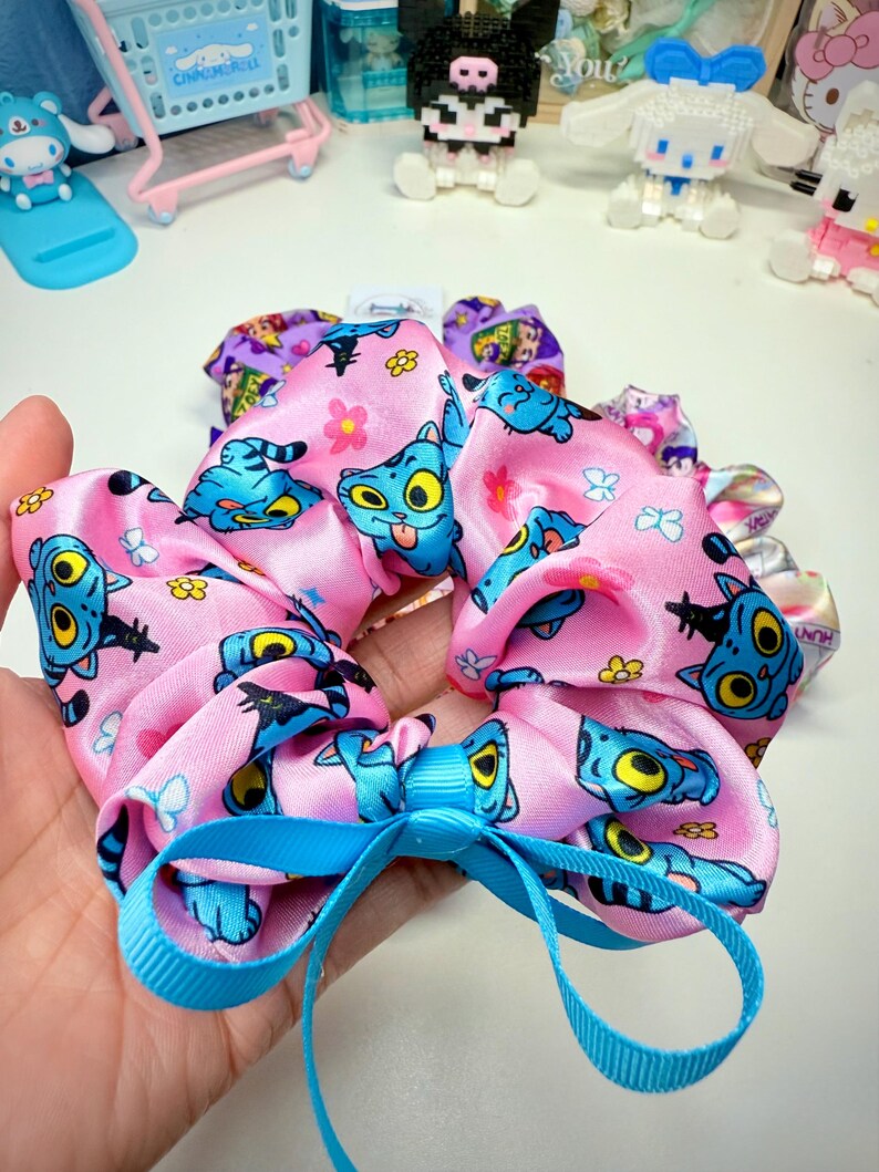 May include: Pink satin scrunchie with a repeating cartoon cat pattern in blue and yellow. A light blue ribbon is tied in a bow at the center. Other scrunchies with different patterns are visible in the background.