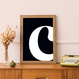 以下が含まれることがあります： 太字でスタイリッシュなフォントで書かれた「C」の文字の白黒の額装プリント。プリントは、木製のドレッサーの上にヴィンテージのタイプライターが置かれた薄いピンクの壁にかけられています。