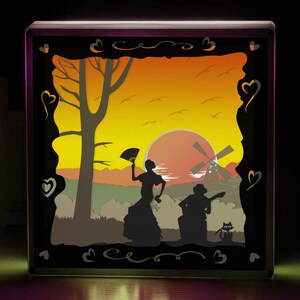 Peut inclure: Boîte lumineuse décorative avec une scène de silhouette en couches. Le design comprend un coucher de soleil, un moulin à vent, un arbre, des personnages et un chat. Le cadre noir est orné de découpes en forme de cœur et de volutes.
