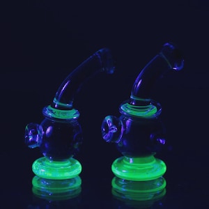 May include: Two glass smoking pipes with a neon green base and accents. The pipes have a curved neck and a bulbous body. The glass is clear with a dark blue hue, and the neon green parts glow brightly.