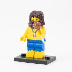 Puede incluir: Una minifigura de Lego de un hombre con cabello castaño y barba, vistiendo una camiseta amarilla y pantalones cortos azules. Sostiene un objeto transparente y amarillo con una esfera roja. Un collar de concha cuelga de su cuello. La figura está sobre una base negra.