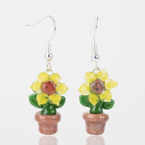 May include: A pair of dangle earrings featuring a sunflower design. Each earring has a yellow flower with a brown center, green leaves, and a brown pot. The earrings are suspended from silver-colored hooks.