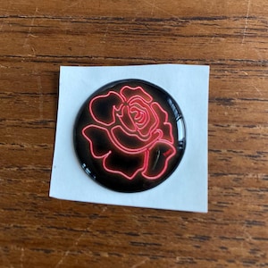 May include: A round, black sticker with a neon red rose design. The rose is outlined with a bright red glow, creating a striking contrast against the black background. The sticker is on a white, rectangular piece of paper.