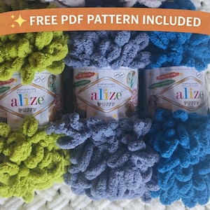 Puede incluir: Tres ovillos de hilo Alize Puffy en tonos verdes, azules y celeste. El hilo es esponjoso y texturizado, con la inscripción "FREE PDF PATTERN INCLUDED" en una pancarta en la parte superior. El hilo está diseñado para proyectos sin aguja ni ganchillo.