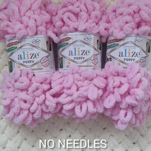 May include: Three spools of Alize Puffy yarn in a soft pink hue. The yarn is displayed with the labels visible, and the text "NO NEEDLES" is at the bottom. The yarn is designed for no-needle knitting projects.