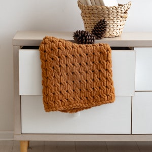 Op de afbeelding: Een witte commode met drie lades en bruine houten poten. Een bruine gebreide deken is over de bovenste lade gelegd. Een rieten mand met boeken en dennenappels staat op de commode.