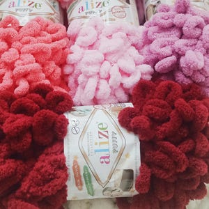 Op de afbeelding: Close-up van verschillende strengen Alize Puffy garen in verschillende tinten roze en rood. Het garen is zacht en getextureerd, met een lusontwerp. De verpakking bevat de tekst "Alize Puffy" en "No Needles No Hooks!"