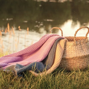 Pode incluir: Uma cesta de vime com um cobertor de malha rosa, azul e bege drapeado sobre ela. A cesta e o cobertor est&atilde;o sentados na grama verde em frente a um corpo de &aacute;gua.