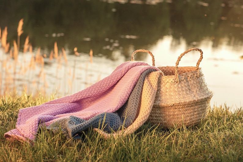Pode incluir: Uma cesta de vime com um cobertor de malha rosa, azul e bege drapeado sobre ela. A cesta e o cobertor est&atilde;o sentados na grama verde em frente a um corpo de &aacute;gua.