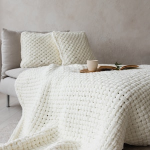 Könnte beinhalten: Eine weiße gestrickte Wolldecke, die über ein Sofa gelegt ist, mit zwei weißen gestrickten Kissen. Die Decke hat eine grobmaschige Strickstruktur und ist über die Kante des Sofas gelegt. Eine kleine Tasse und Untertasse sowie ein Buch liegen auf dem Sofa.