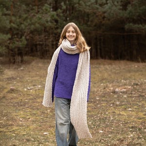 May include: A person wearing a purple sweater, blue jeans, and a cream-colored chunky knit scarf. The person is walking outdoors in a natural setting with trees in the background.