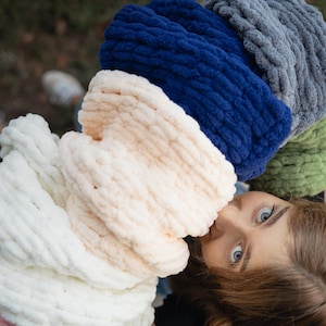 May include: A stack of colourful chunky knit yarn in shades of pink, white, cream, green, blue and grey. The yarn is arranged in a circle around a person's head.