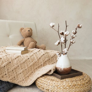 Pode incluir: Uma poltrona de couro branco com um urso de peluche castanho sentado nela. Uma manta de malha está drapeada sobre a poltrona. Um vaso com ramos de algodão está sobre um pufe tecido.