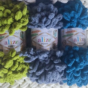 Puede incluir: Tres ovillos de lana Alize Puffy en tonos verde, azul y turquesa. La lana es gruesa y en bucles, diseñada para proyectos sin aguja ni ganchillo. La colección Alize Puffy se muestra sobre una superficie blanca texturizada.