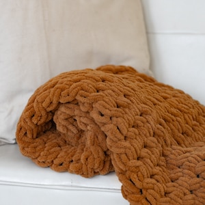 May include: A brown chunky knit throw blanket draped over a white couch with a beige pillow.