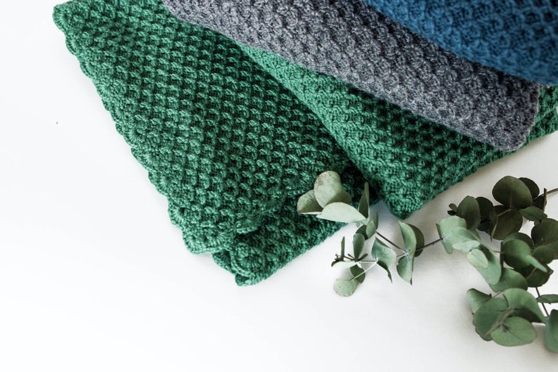 Pode incluir: Uma pilha de cobertores de malha nas cores verde, cinza e azul. Um ramo de folhas de eucalipto &eacute; colocado na frente dos cobertores.