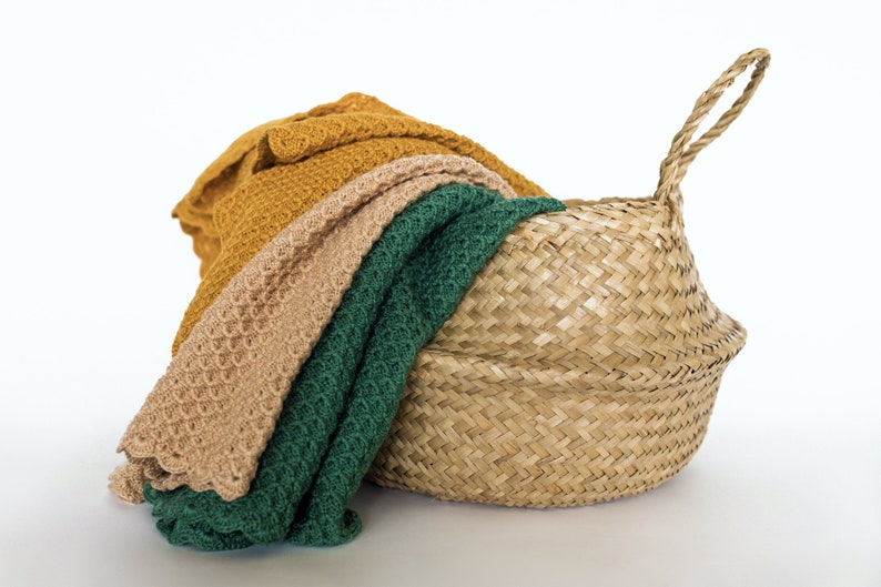 Pode incluir: Uma cesta de vime tecida com al&ccedil;a, cheia de tr&ecirc;s cobertores de malha em tons de verde, bege e amarelo mostarda.