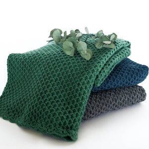 Pode incluir: Uma pilha de tr&ecirc;s cobertores de malha em verde, azul e cinza. O cobertor superior &eacute; verde e tem uma textura de waffle. Um ramo de folhas de eucalipto &eacute; colocado em cima dos cobertores.