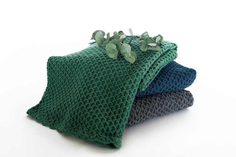Pode incluir: Uma pilha de tr&ecirc;s cobertores de malha em verde, azul e cinza. O cobertor superior &eacute; verde e tem uma textura de waffle. Um ramo de folhas de eucalipto &eacute; colocado em cima dos cobertores.