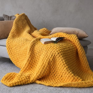 Op de afbeelding: Een gele gebreide deken gedrapeerd over een grijze bank met kussens. Een boek ligt op de deken.