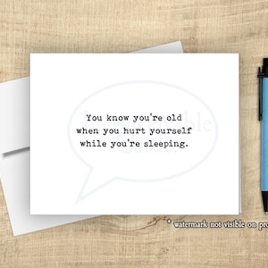 May include: A white greeting card with the text "You know you're old when you hurt yourself while you're sleeping" in a speech bubble. A blue pen rests on a wooden surface next to the card and a white envelope.