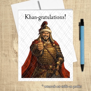 May include: Greeting card with the text "Khan-gratulations!" and an illustration of a smiling warrior giving a thumbs-up. The warrior is wearing ornate armor and a red cape. A blue pen rests beside the card.