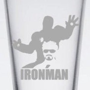 Puede incluir: Un vaso de pinta de vidrio transparente con una silueta gris de Iron Man y el texto "IRONMAN" impreso en la parte inferior.