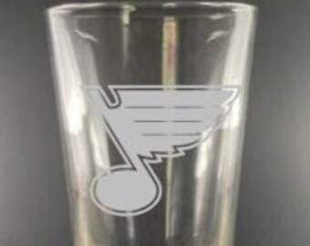 May include: A clear glass drinking glass with a white etched design of the St. Louis Blues hockey team logo. The logo is a stylized note with a wing on top.