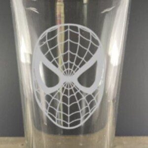 Puede incluir: Un vaso de pinta de vidrio transparente con un dise&ntilde;o grabado en blanco de la m&aacute;scara de Spider-Man.