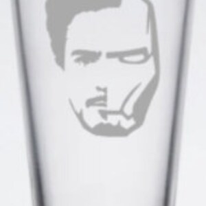 Puede incluir: Un vaso de pinta de vidrio transparente con una silueta blanca de la cara de Iron Man grabada en la parte delantera.