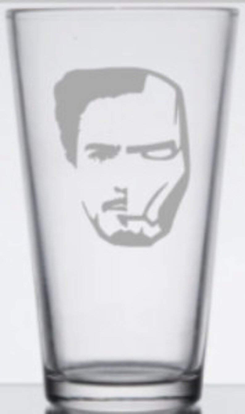 Puede incluir: Un vaso de pinta de vidrio transparente con una silueta blanca de la cara de Iron Man grabada en la parte delantera.