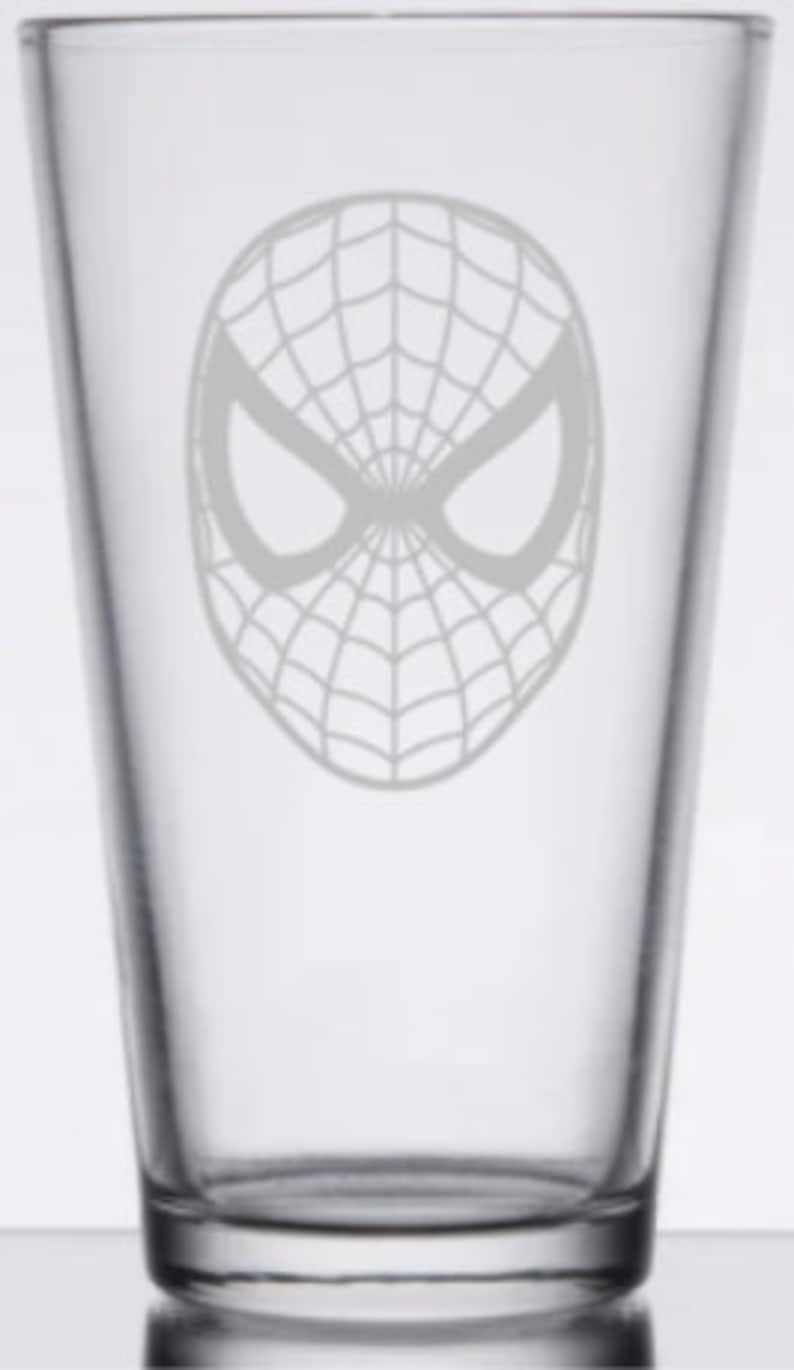 Puede incluir: Un vaso de pinta de vidrio transparente con un dise&ntilde;o grabado en blanco de la m&aacute;scara de Spider-Man.