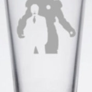 Puede incluir: Un vaso de pinta de vidrio transparente con un dise&ntilde;o grabado en blanco de Iron Man en su traje y Tony Stark en traje de pie detr&aacute;s de &eacute;l.