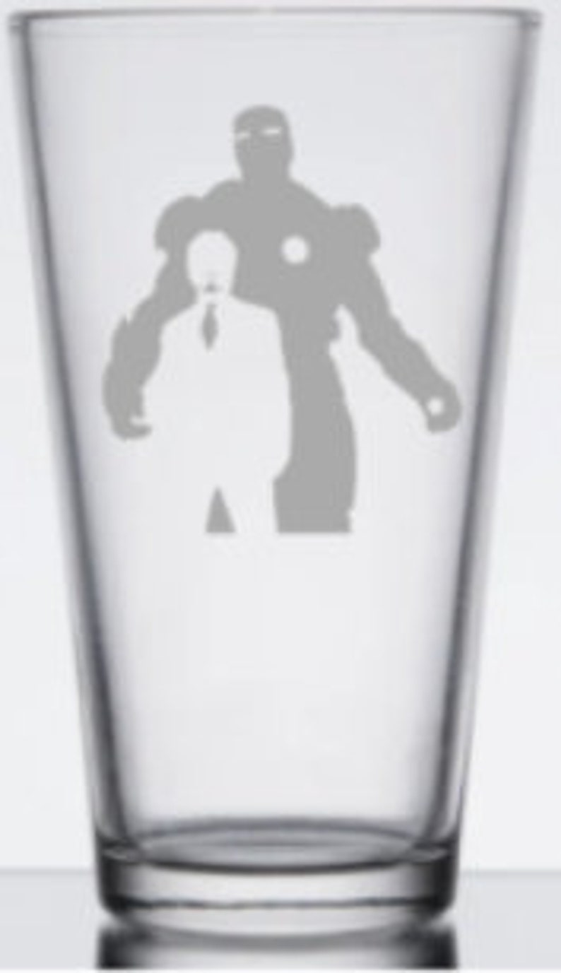 Puede incluir: Un vaso de pinta de vidrio transparente con un dise&ntilde;o grabado en blanco de Iron Man en su traje y Tony Stark en traje de pie detr&aacute;s de &eacute;l.