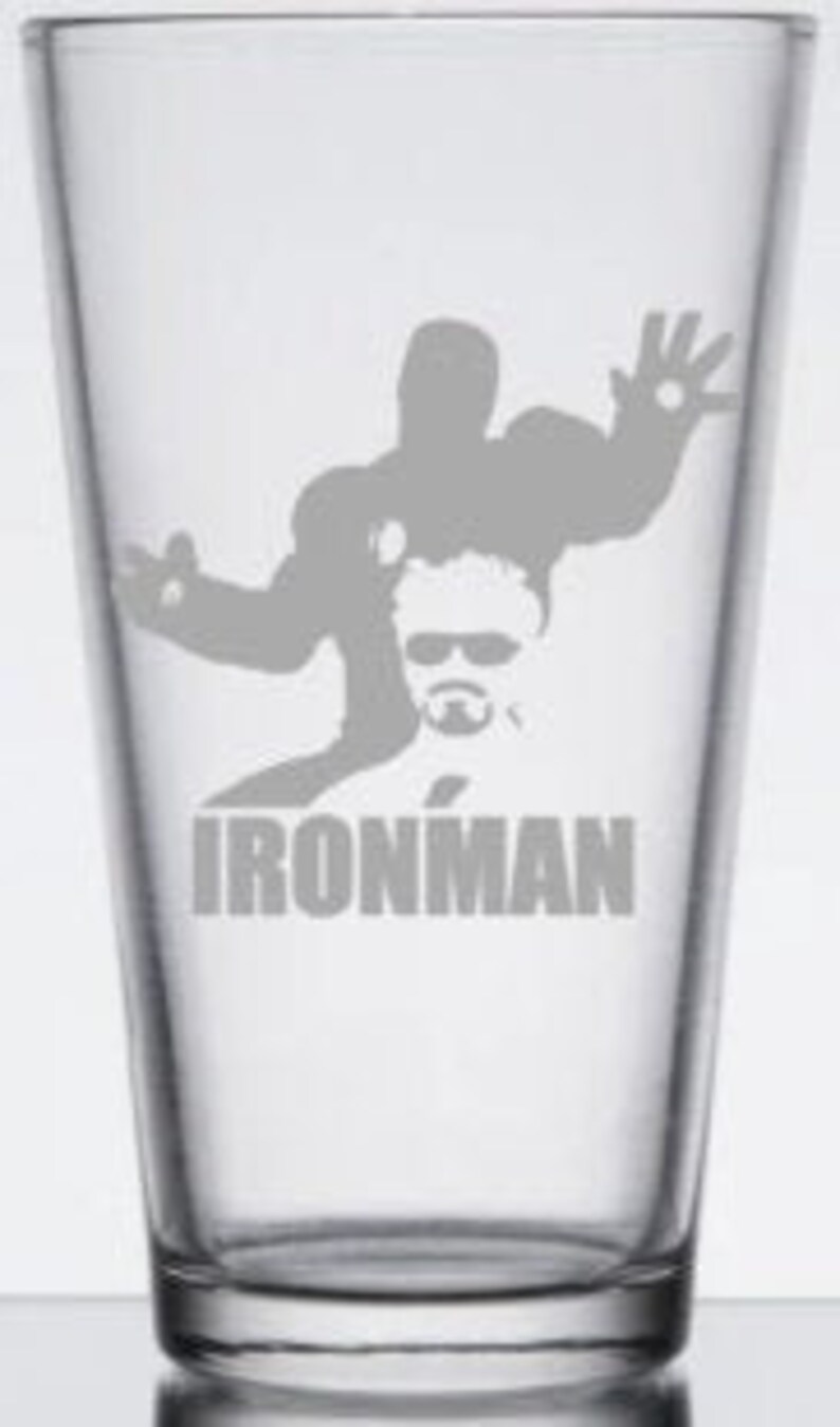 Puede incluir: Un vaso de pinta de vidrio transparente con una silueta gris de Iron Man y el texto "IRONMAN" impreso en la parte inferior.