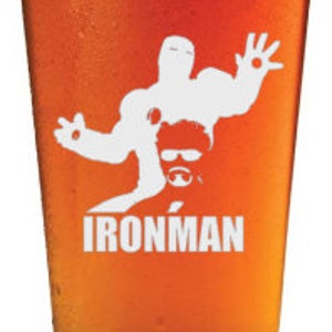 Puede incluir: Un vaso de pinta de vidrio transparente con una silueta blanca de Iron Man impresa en el lateral. El vaso est&aacute; lleno de una cerveza de color &aacute;mbar claro.