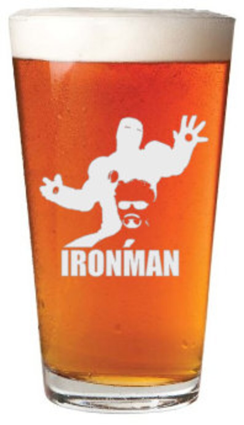 Puede incluir: Un vaso de pinta de vidrio transparente con una silueta blanca de Iron Man impresa en el lateral. El vaso est&aacute; lleno de una cerveza de color &aacute;mbar claro.