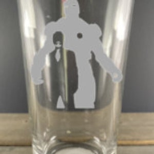 Puede incluir: Un vaso de pinta de vidrio transparente con una silueta blanca de Iron Man y otra figura de pie detr&aacute;s de &eacute;l.